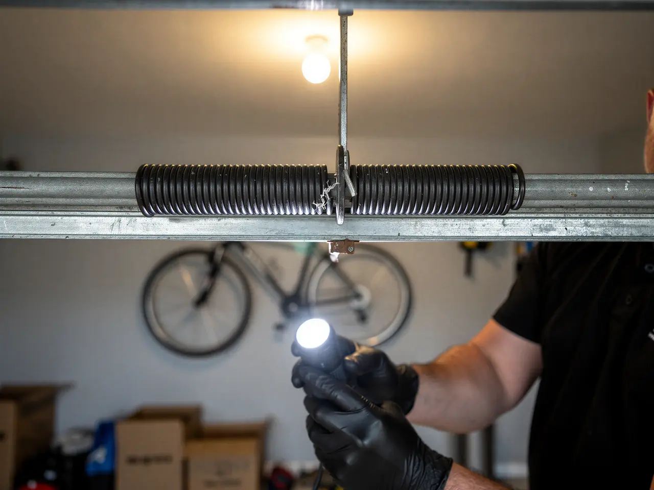 A technician in black gloves shines a flashlight on a garage door torsion spring.